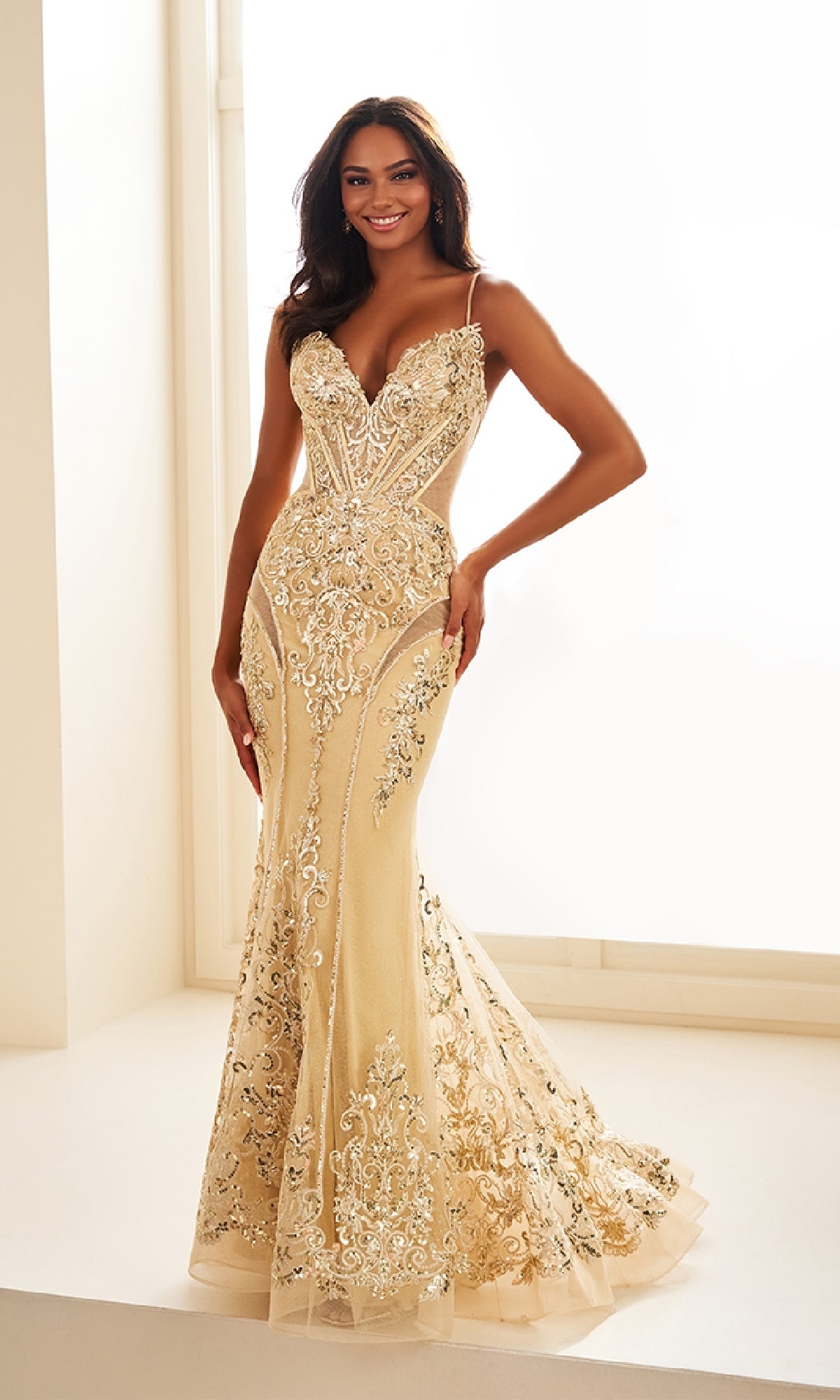 A woman poses indoors, smiling in the Ellie Wilde EW36006 long prom dress—an elegant, fitted gold embellished mermaid gown with spaghetti straps and intricate lace detailing that shimmers with flair.